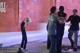 A barefoot person in a black top raises a white board toward a masked figure as three men approach near a pink-tiled wall with 'ART JOG' on the left.