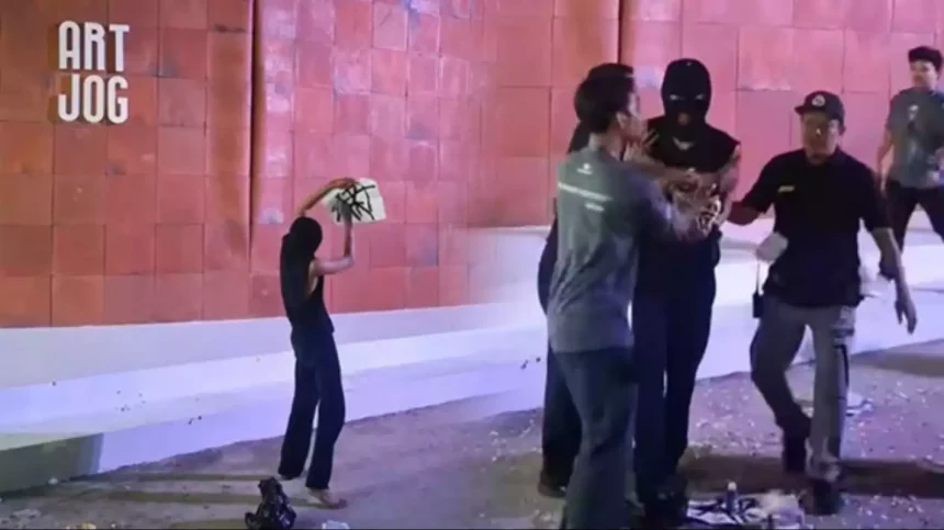 A barefoot person in a black top raises a white board toward a masked figure as three men approach near a pink-tiled wall with 'ART JOG' on the left.