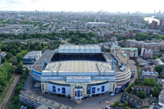 Aerial view of a large blue-and-white stadium surrounded by trees, streets, and dense city housing.