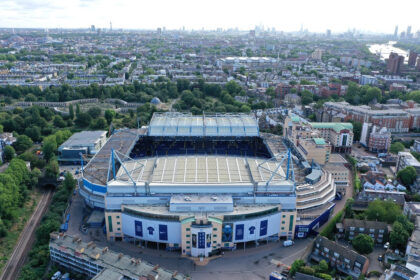 Aerial view of a large blue-and-white stadium surrounded by trees, streets, and dense city housing.