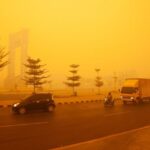 City street shrouded in thick orange smog with cars, motorcycles, and a truck visible on the road.