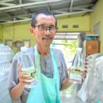 Middle-aged man with glasses and a teal apron holding two bottles of water in a warehouse setting.