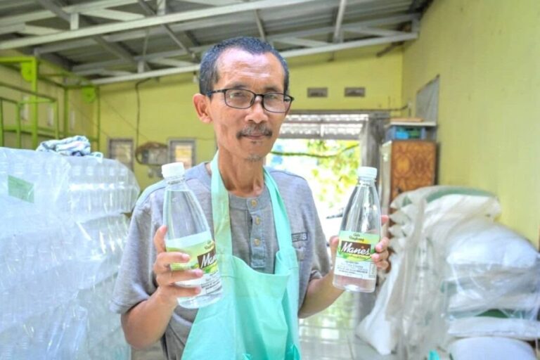 Middle-aged man with glasses and a teal apron holding two bottles of water in a warehouse setting.