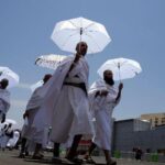 Group of men in white robes walking in a street under a bright blue sky, carrying white parasols as part of a daytime procession.