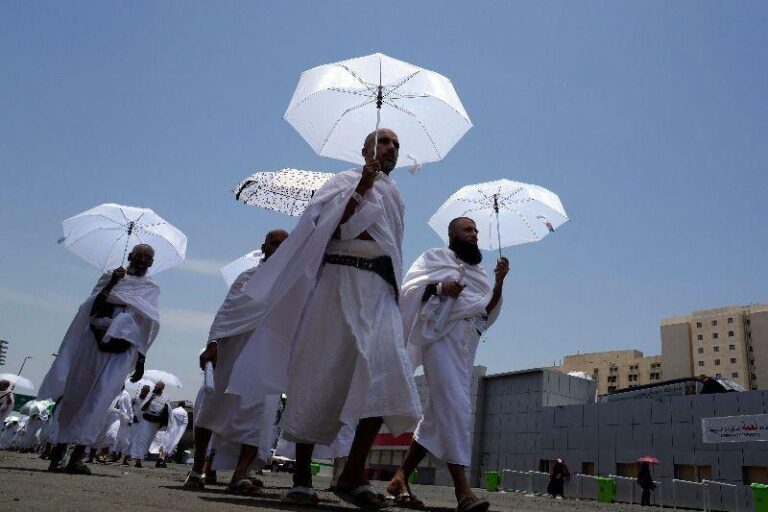 Group of men in white robes walking in a street under a bright blue sky, carrying white parasols as part of a daytime procession.