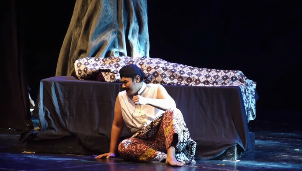 suluk kang pungkasan-ezremove - Mabur.co Performer sitting on the stage floor in colorful patterned pants and a white sleeveless shirt, gesturing with one hand near a covered prop table.