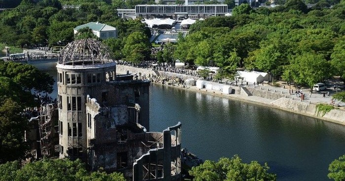 Ruined circular church tower by a river, surrounded by trees and a riverside walkway with visitors