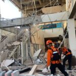 Rescue workers in orange uniforms and helmets navigate a collapsed building amid tangled metal and rubble.