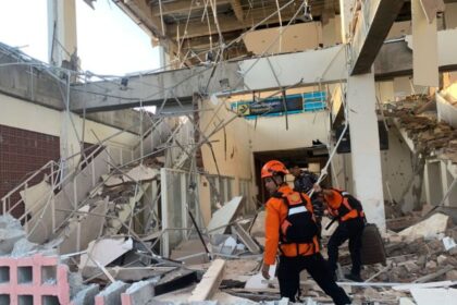 Rescue workers in orange uniforms and helmets navigate a collapsed building amid tangled metal and rubble.