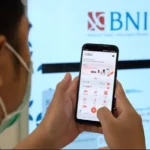 Close-up of a person wearing a mask holding a smartphone displaying the BNI mobile app, with a large BNI logo on a screen in the background.