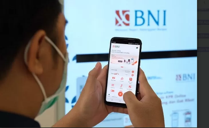 Close-up of a person wearing a mask holding a smartphone displaying the BNI mobile app, with a large BNI logo on a screen in the background.