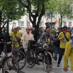 Group of people with bicycles gathered in a city square, some wearing yellow shirts and hats, taking photos and chatting.