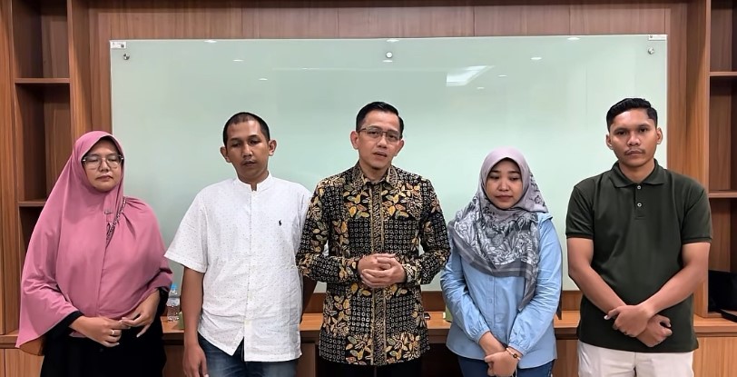Five adults stand in a row in an office with a glass whiteboard behind them, posing for a group photo.