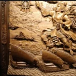 Man in patterned shirt stands in profile, viewing a large carved wooden relief of a soaring warrior goddess and ships below.