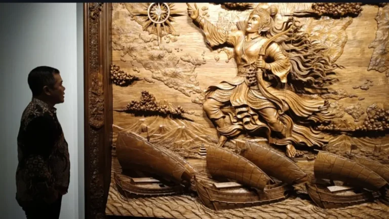 Man in patterned shirt stands in profile, viewing a large carved wooden relief of a soaring warrior goddess and ships below.
