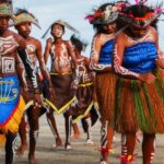 Dancers in colorful traditional costumes with body paint perform on a sandy beach.