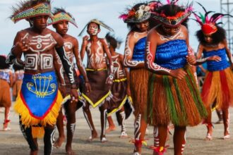 Dancers in colorful traditional costumes with body paint perform on a sandy beach.
