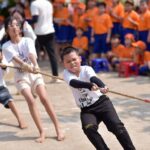 Group of kids in white shirts tugging on a rope during a school event, with a crowd of orange-shirted spectators in the background.