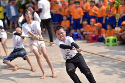 Group of kids in white shirts tugging on a rope during a school event, with a crowd of orange-shirted spectators in the background.