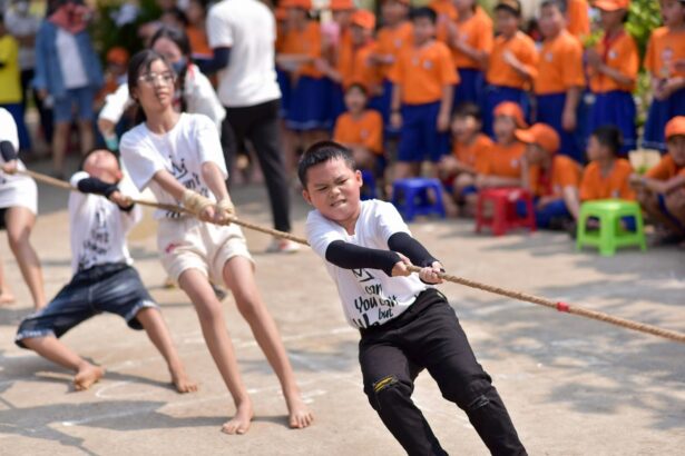 Group of kids in white shirts tugging on a rope during a school event, with a crowd of orange-shirted spectators in the background.