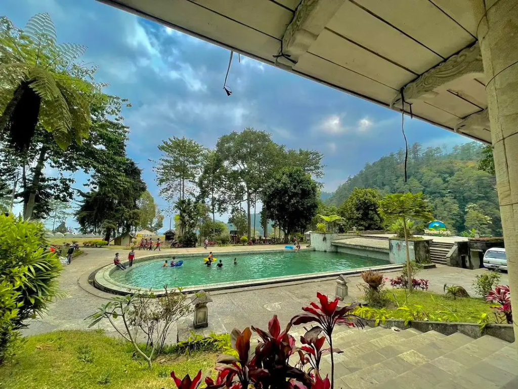 Outdoor pool at a resort, surrounded by trees and hills, with people swimming and relaxing on the deck.
