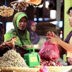 Women at a busy market stall choose and bag produce; a green scale sits on the counter nearby.