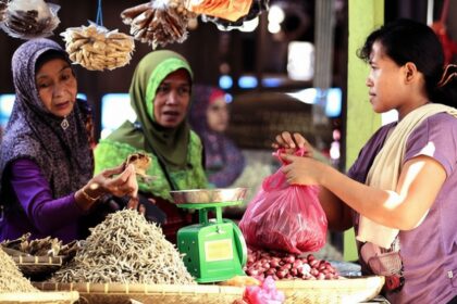 Women at a busy market stall choose and bag produce; a green scale sits on the counter nearby.