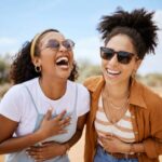 Two women laughing together outdoors, wearing sunglasses, enjoying a sunny day in a dry, open landscape.