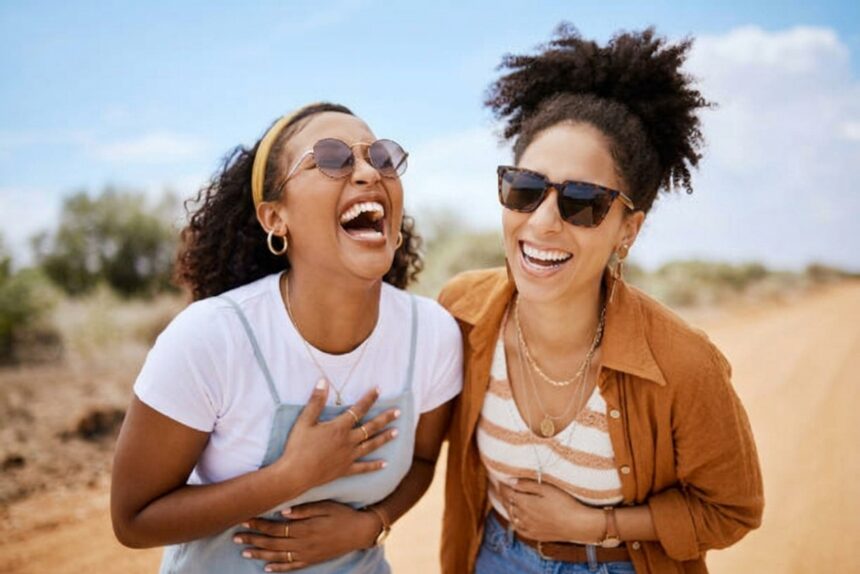 Two women laughing together outdoors, wearing sunglasses, enjoying a sunny day in a dry, open landscape.