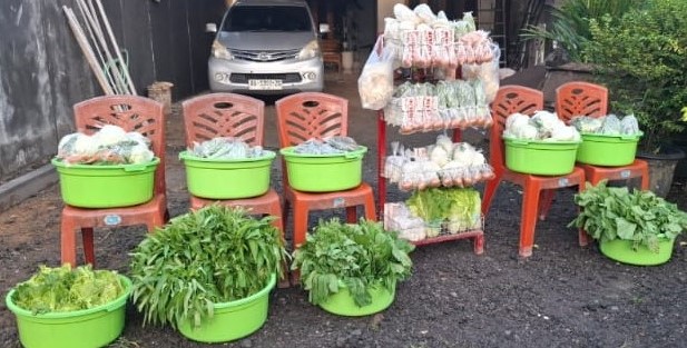 tgdfg - Mabur.co Street market stall with green buckets of leafy greens placed on orange plastic chairs and stacked snack bags in front of a parked car.