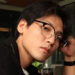 Young man with round glasses and dark hair resting his head on his hand in a modern cafe, looking at the camera.