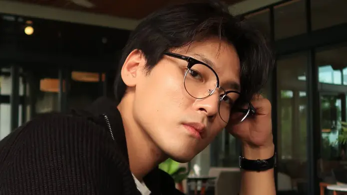 Young man with round glasses and dark hair resting his head on his hand in a modern cafe, looking at the camera.