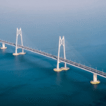 Suspension bridge spanning a calm blue ocean, featuring tall white towers and curved approach leads over the water.