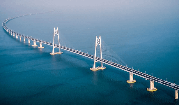 Suspension bridge spanning a calm blue ocean, featuring tall white towers and curved approach leads over the water.