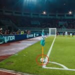 Soccer corner; a ball rests near the flag at the corner of the field with players and spectators in the background.