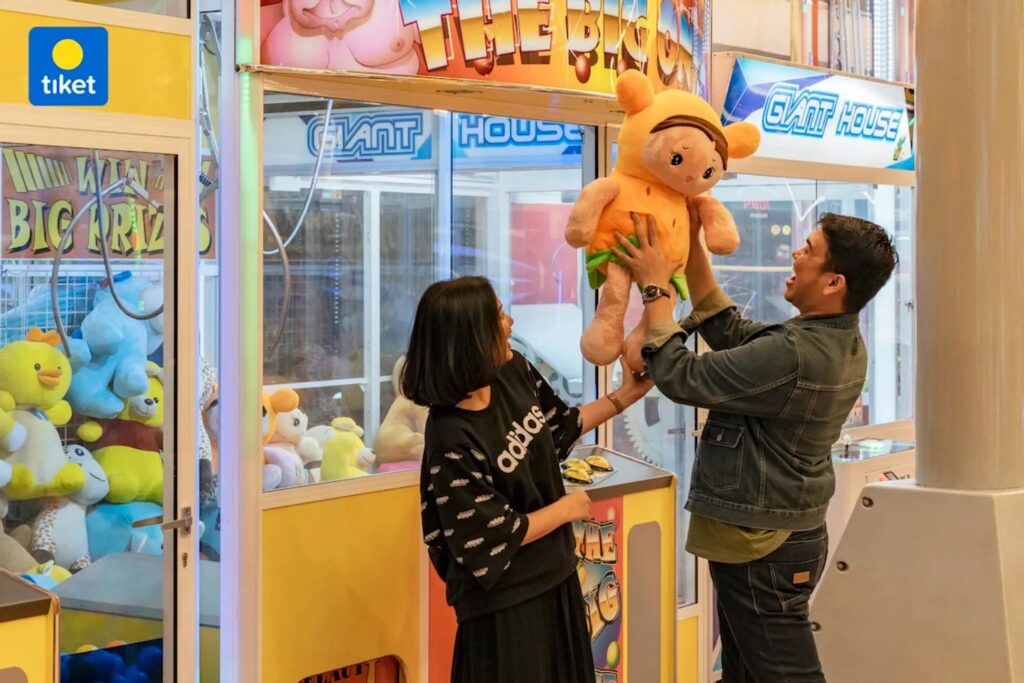 A man hands a large orange plush toy to a smiling woman inside a bright arcade claw machine area, with stuffed animals visible inside the machine.