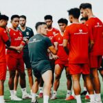 Soccer team in red uniforms forming a huddle on a green field as the coach explains a tactic to them.