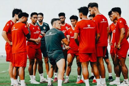 Soccer team in red uniforms forming a huddle on a green field as the coach explains a tactic to them.