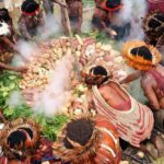 Indigenous people in feather headdresses and body paint gather around a mound of root vegetables while smoke rises from a central fire.