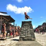 A performer leaps from a stone pedestal in a sunny village square, surrounded by people in traditional dress watching the act.