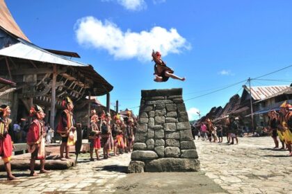 A performer leaps from a stone pedestal in a sunny village square, surrounded by people in traditional dress watching the act.