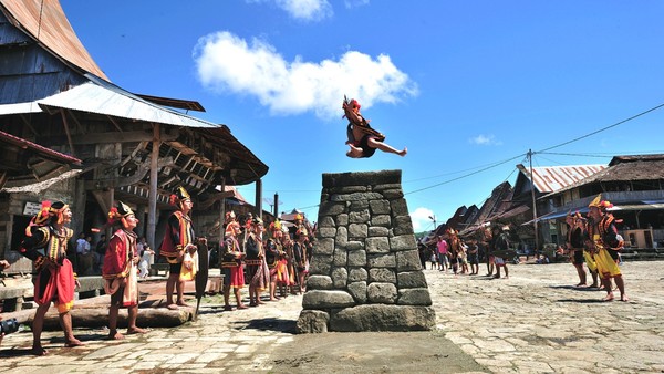 A performer leaps from a stone pedestal in a sunny village square, surrounded by people in traditional dress watching the act.