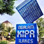 Public outdoor art installation: a blue cube covered in white binary digits balanced on its edge, resting above a blue pedestal with bold white typography, in a plaza with trees and buildings in the background.