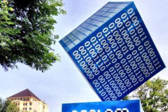 Public outdoor art installation: a blue cube covered in white binary digits balanced on its edge, resting above a blue pedestal with bold white typography, in a plaza with trees and buildings in the background.