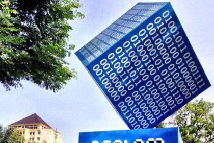 Public outdoor art installation: a blue cube covered in white binary digits balanced on its edge, resting above a blue pedestal with bold white typography, in a plaza with trees and buildings in the background.