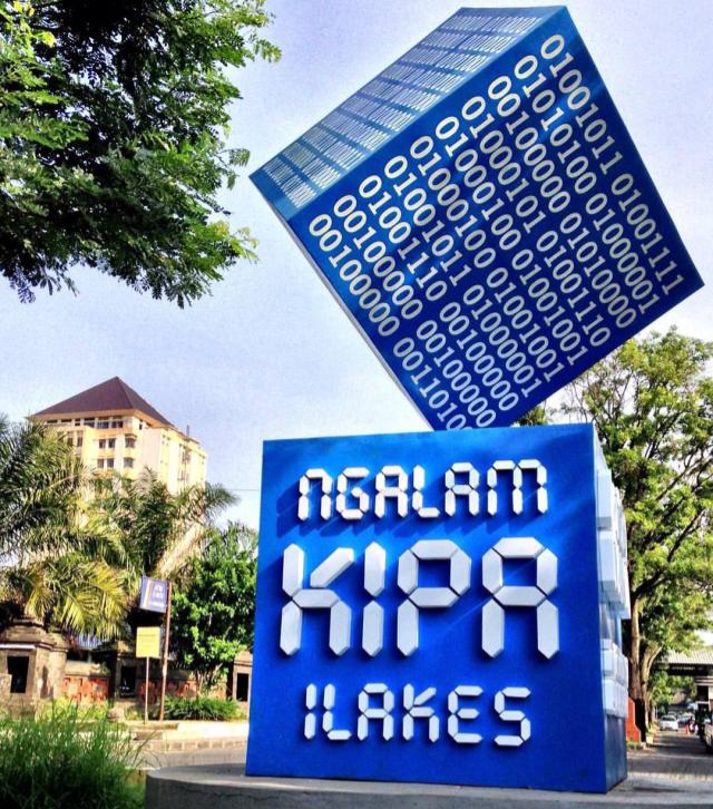 Public outdoor art installation: a blue cube covered in white binary digits balanced on its edge, resting above a blue pedestal with bold white typography, in a plaza with trees and buildings in the background.