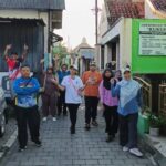 Group of people walking down a narrow street, waving at the camera, on a sunny day.