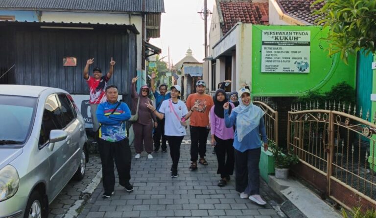 Group of people walking down a narrow street, waving at the camera, on a sunny day.
