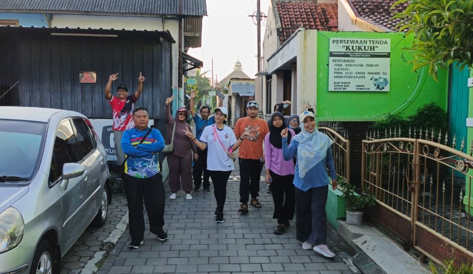 Group of people walking down a narrow street, waving at the camera, on a sunny day.