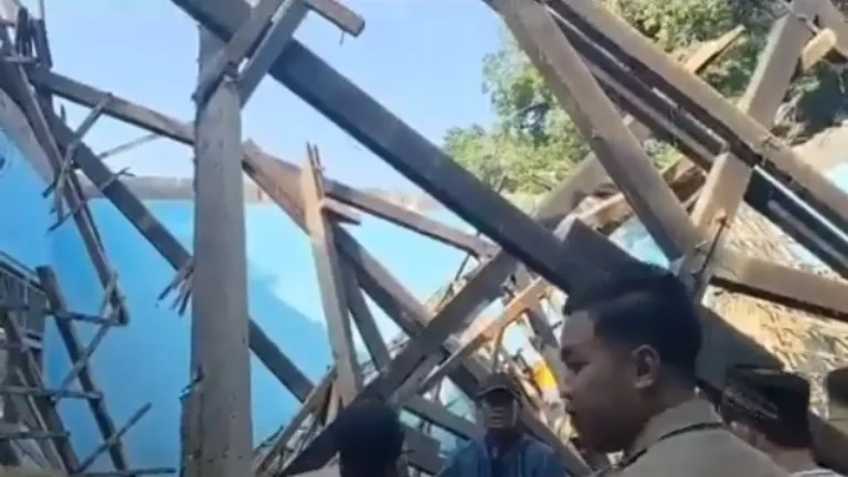 Group of people standing amid a collapsed timber frame with diagonal beams and a blue exterior wall behind them, inspecting the wreckage.